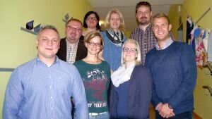 Gruppenfoto von acht Personen, die nebeneinander in einem Flur stehen und in die Kamera lächeln. Die Personen tragen Freizeit- und Businesskleidung und stehen vor einer gelblich gestrichenen Wand mit einigen dekorativen Elementen im Hintergrund.