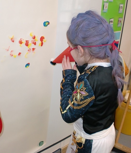 Foto, auf dem ein Mädchen mit lila Perücke von hinten zu sehen ist. Sie malt mit einer aus Papier gebastelten Nase in Trichterform, die sie am Kopf befestigt hat, mit Fingermalfarben auf ein großes Papier, das an der Wand befestigt ist.