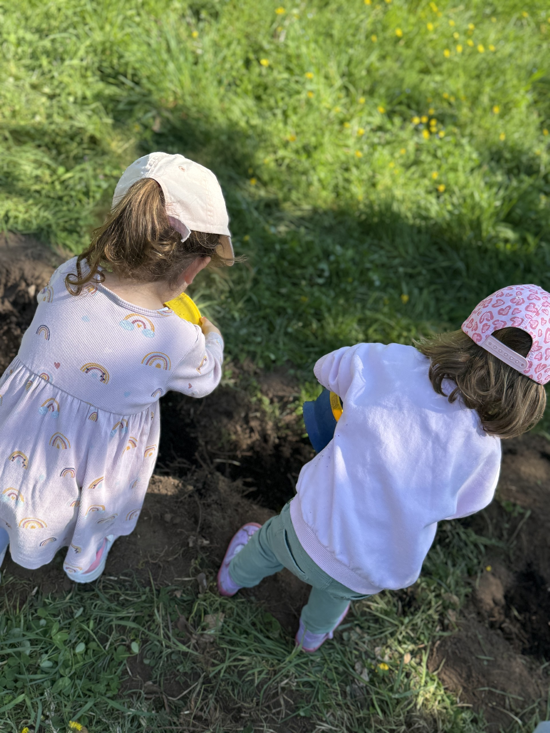 Zwei Kinder sind von hinten zu sehen, die auf dem Boden kniend Pflanzen in Erde einbuddeln