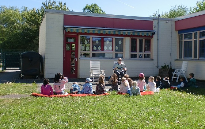 Eine Gruppe Kinder sitzt im Gras und hört einer erwachsenen Person zu, die vor ihnen auf einem Stuhl sitzt und ein Buch vorliest