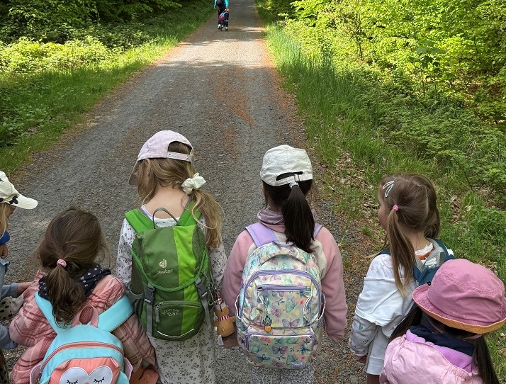 Kindergruppe läuft auf einem Waldweg entlang, weit vorne ist eine erwachsene Person mit einem Kind an der Hand zu erkennen.