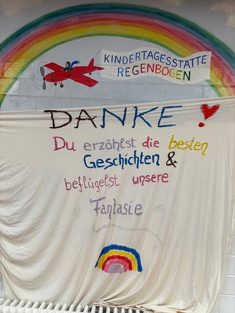 Man sieht die Mauer der Kita Regenbogen. Auf die Mauer ist ein Regenbogen gemalt und ein rotes Flugzeug fliegt darunter, zieht eine Fahne hinter sich her auf der Kita Regenbogen steht.Davor ist ein weißes Bettlaken steht "Danke - du erzählst die besten Geschichten und beflügelst unsere Fantasie"