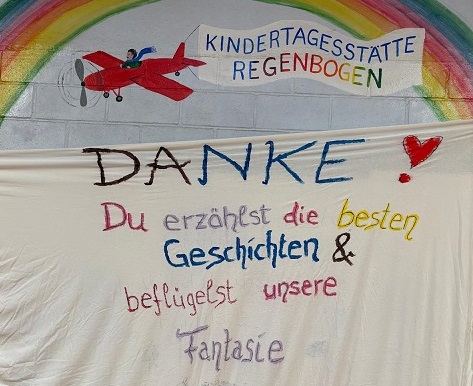 Man sieht die Mauer der Kita Regenbogen. Auf die Mauer ist ein Regenbogen gemalt und ein rotes Flugzeug fliegt darunter, zieht eine Fahne hinter sich her auf der Kita Regenbogen steht.Davor ist ein weißes Bettlaken steht "Danke - du erzählst die besten Geschichten und beflügelst unsere Fantasie"