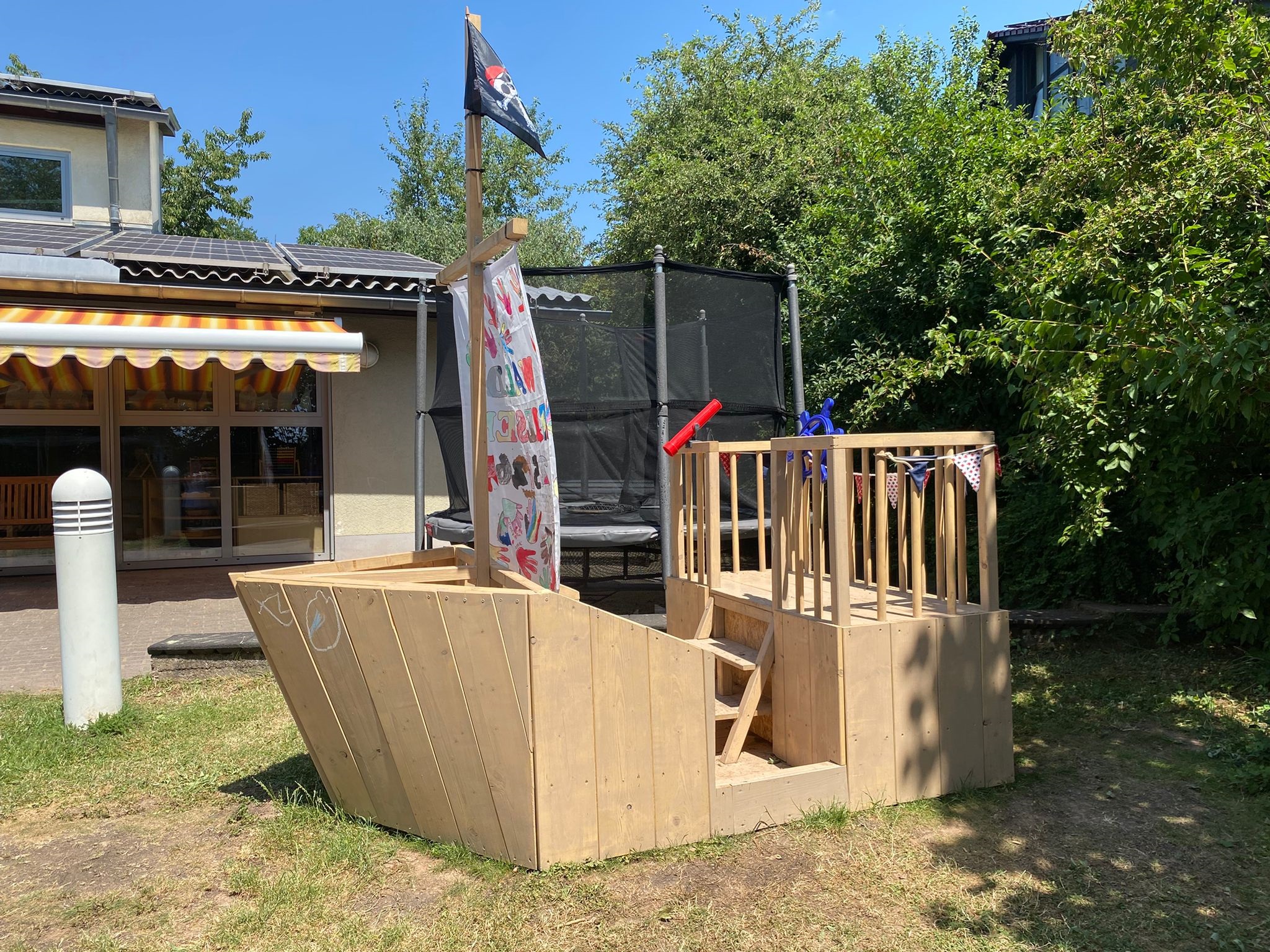 Selbst gebautes Holzschiff, so groß, dass mehrere Kinder darauf spielen können. Auf dem Segel sind bunte Handabdrücke von Kindern
