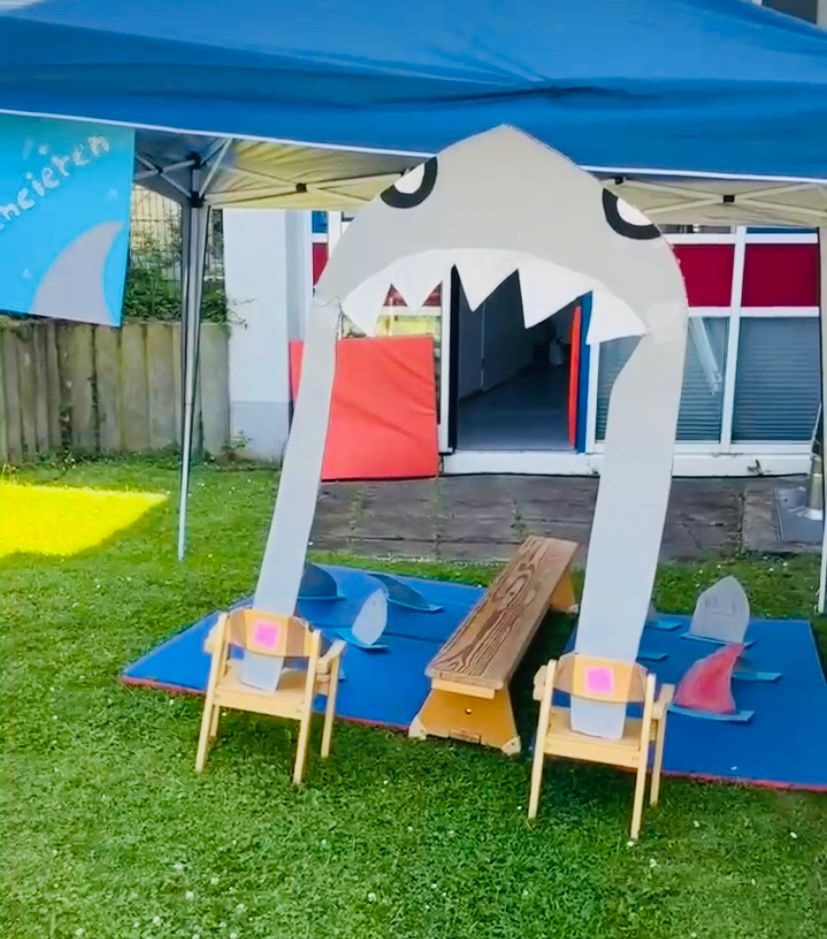 Auf dem Foto sieht man einen aus Pappe gebauten Hai der sein Maul aufreißt, so groß, dass Kinder durchgehen können. Beim Durchgehen müssen sie über eine niedrige Bank laufen.