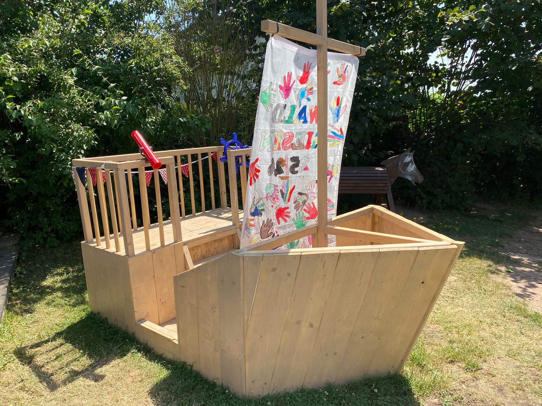 Selbst gebautes Holzschiff, so groß, dass mehrere Kinder darauf spielen können. Auf dem Segel sind bunte Handabdrücke von Kindern