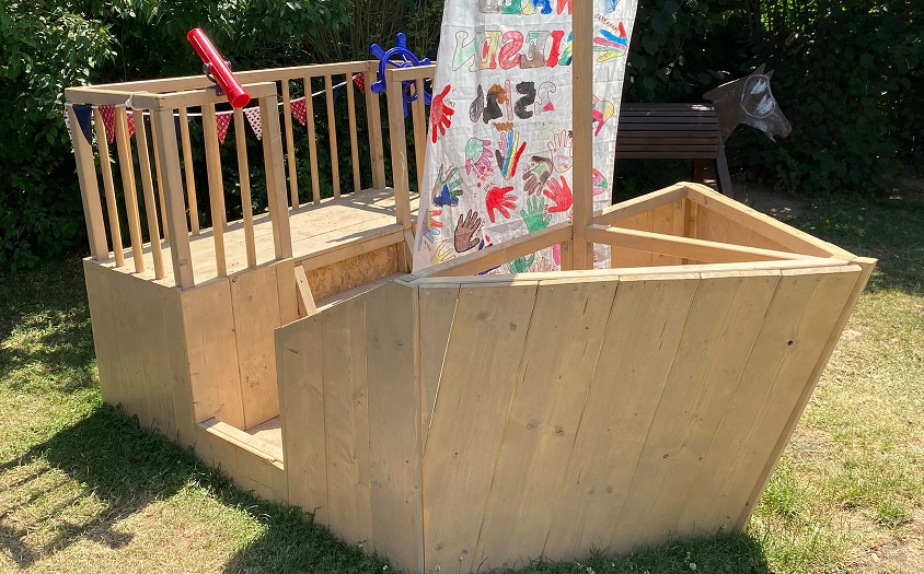 Selbst gebautes Holzschiff, so groß, dass mehrere Kinder darauf spielen können. Auf dem Segel sind bunte Handabdrücke von Kindern