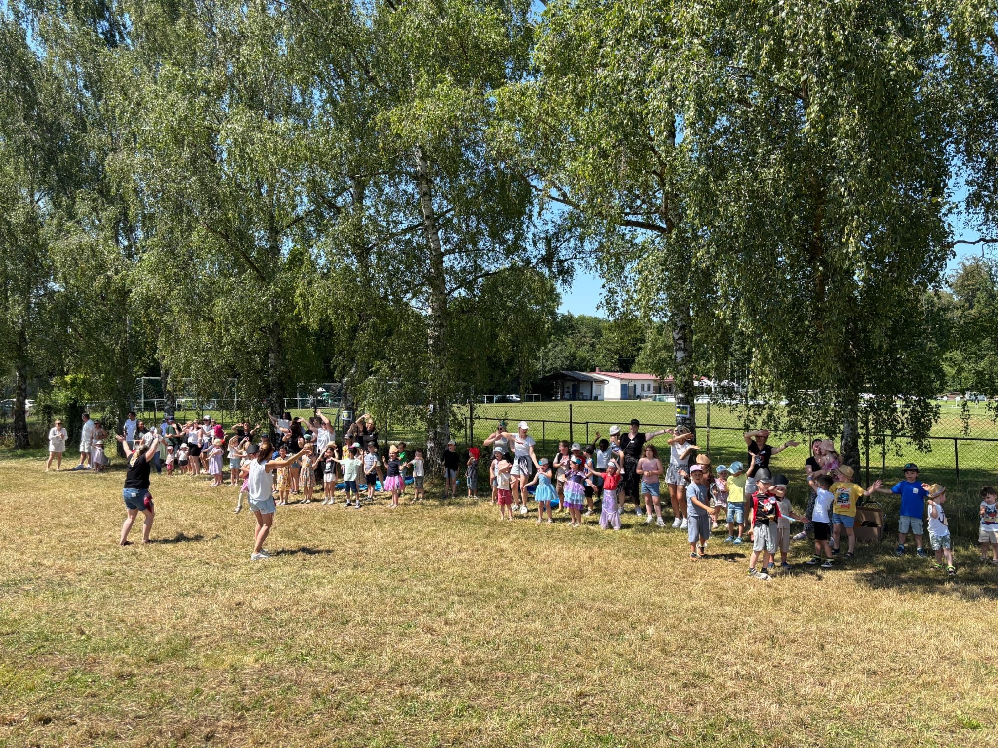 Viele Kinder und ein paar Erwachsene stehen unter Birkenbäumen und führen einen Tanz auf. Zwei Erwachsene stehen gegenüber und tanzen vor.