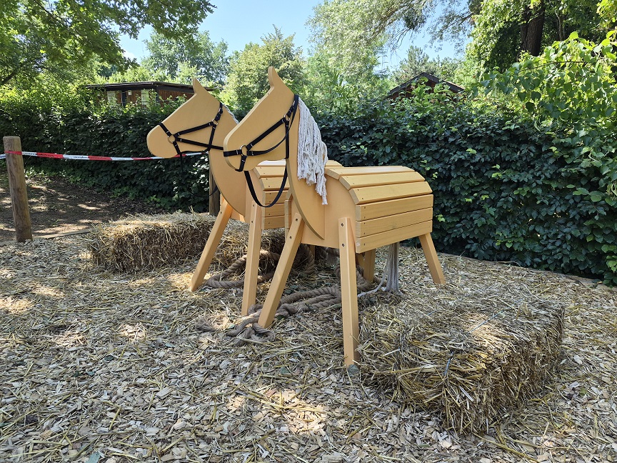 Foto von zwei Holzpferden aus hellem Holz und weißer Mähne aus Wolle die auf Rindenmulch und Stroh stehen im Garten