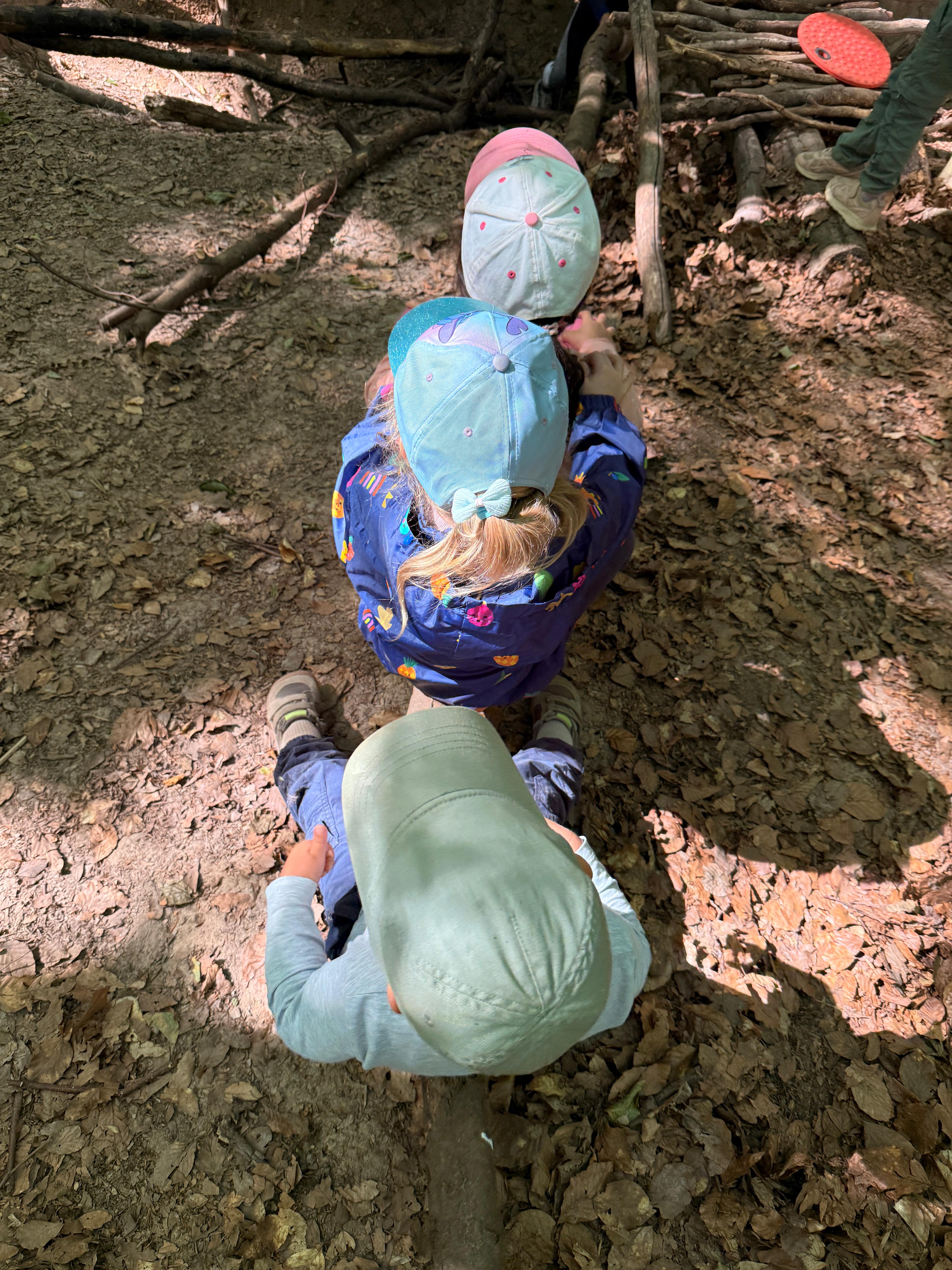 Drei Kinder sind von hinten zu sehen, alle tragen Kappen. Sie sitzen hintereinander auf einem Baumstamm