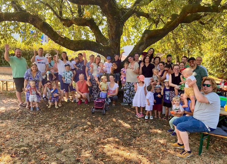 Eine Gruppe von ca 45 Kindern und Erwachsenen steht unter einem verästelten Kirschbaum, einige winken fröhlich in die Kamera